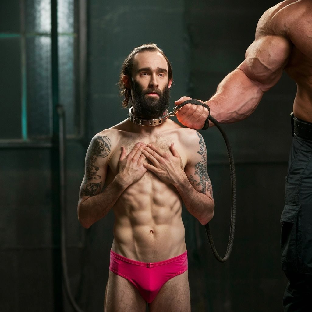 A slim bearded man in pink briefs wearing a collar, his leash held by a muscular arm