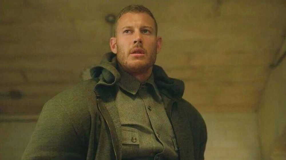 A rugged man in a green military coat looking up in a dimly lit room