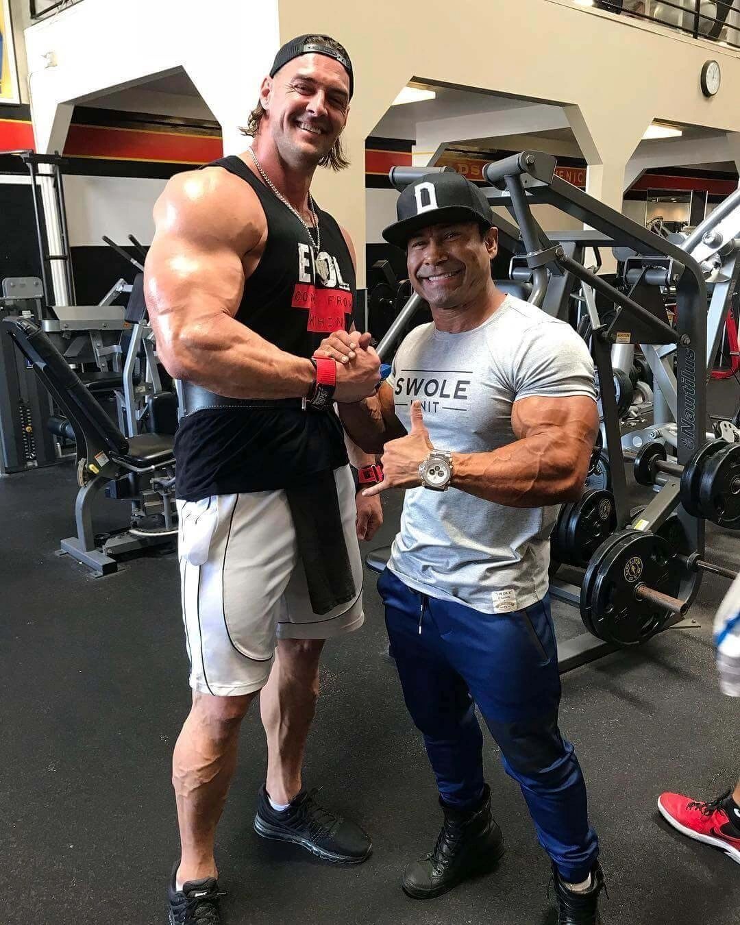 Two massive bodybuilders, one in a vest and one in a cap, posing side by side in a gym