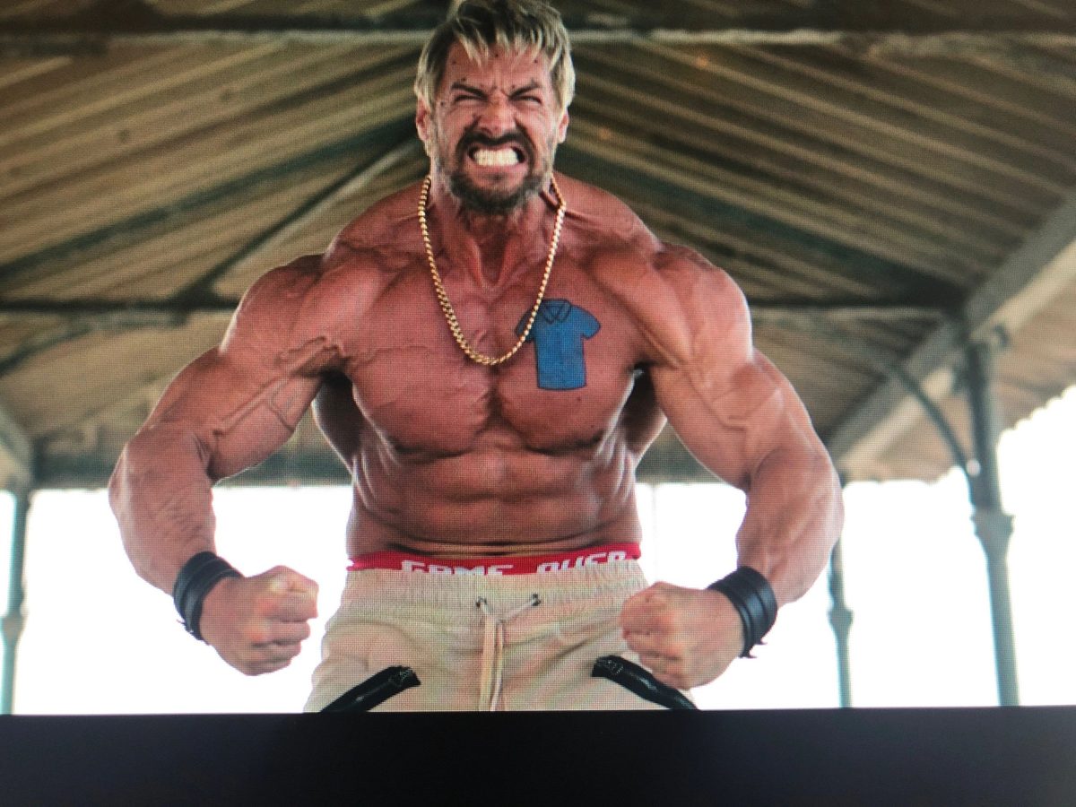 A shirtless bodybuilder with a gold chain snarling and flexing