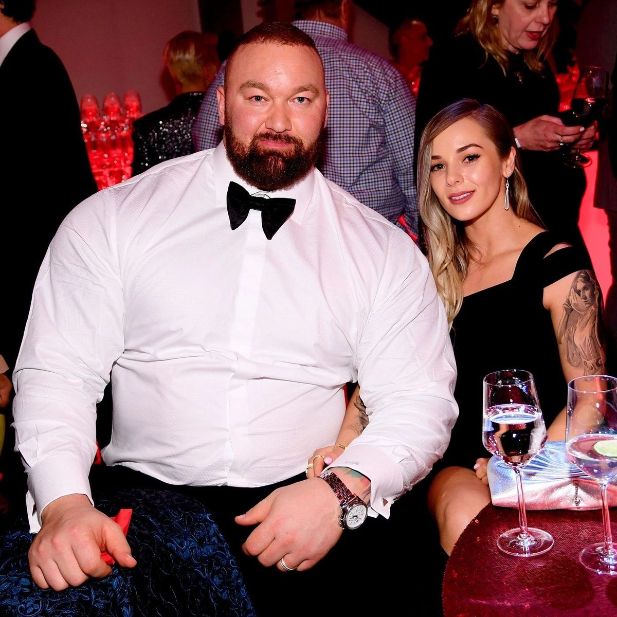 A huge bearded man in a white shirt and bow tie seated beside a woman at an event