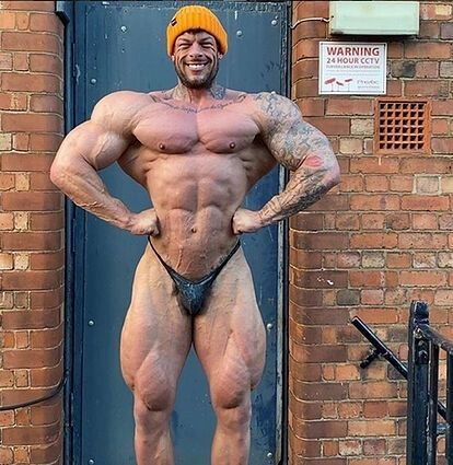 A tattooed bodybuilder in posing trunks and a beanie hitting a lat spread by a brick wall