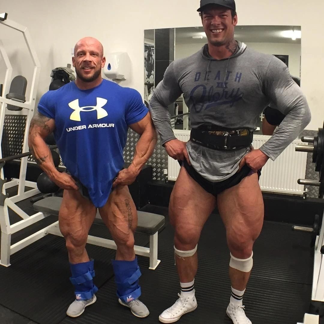Two huge bodybuilders in a gym lifting their tops to show off their massive thighs
