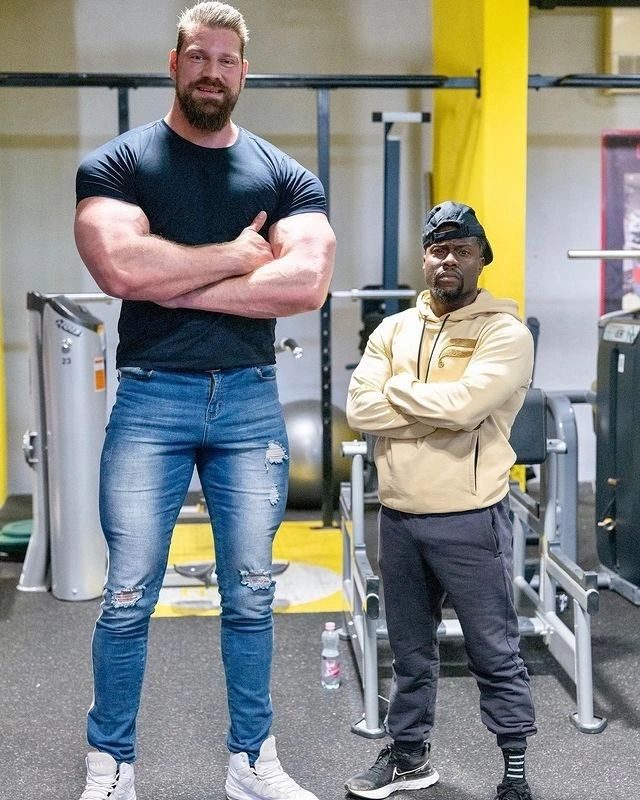 A towering bearded man in a T-shirt and jeans next to a much shorter man in a gym