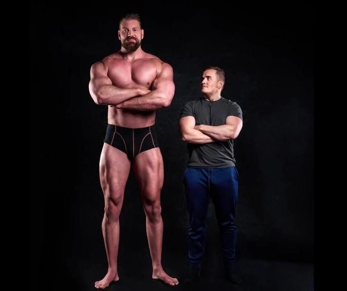 A tall bodybuilder in black trunks standing barefoot beside a shorter man in a T-shirt and jeans