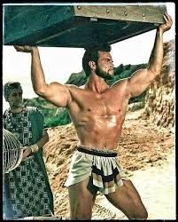 Film still of Steve Reeves in a loincloth lifting a heavy stone slab overhead