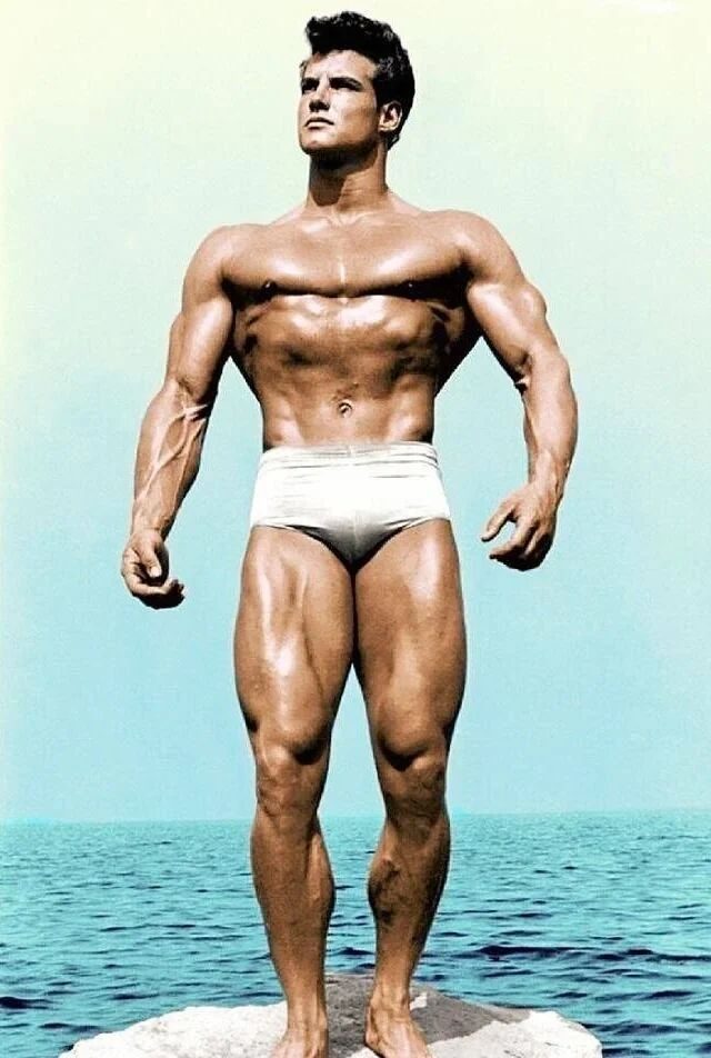Vintage colour photo of Steve Reeves in white trunks standing on a rock by the sea