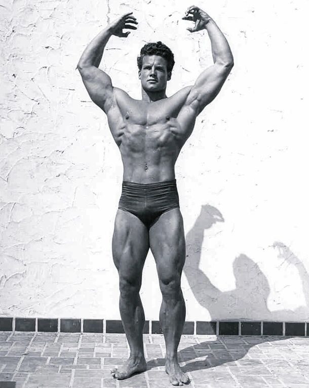 Vintage photo of Steve Reeves in dark trunks flexing against a white wall