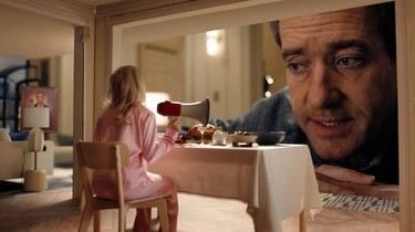 A man peering into a dolls' house room where a tiny woman in pink sits at a table